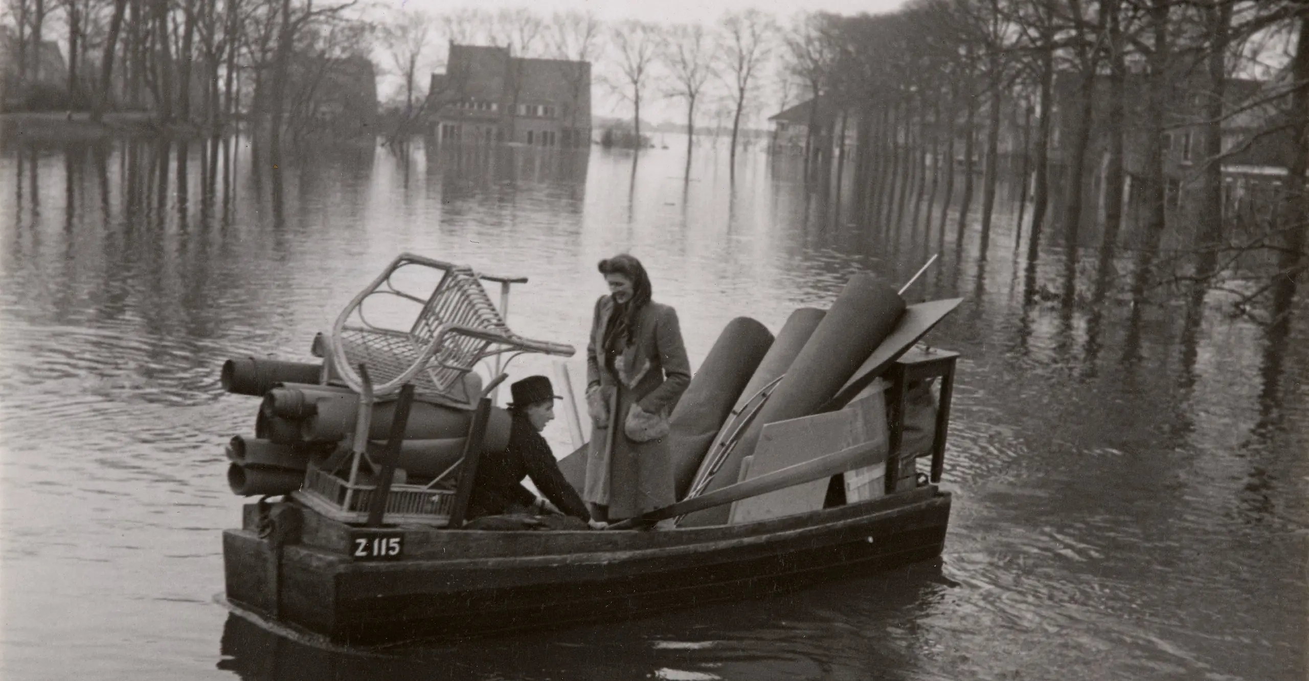 Black and white photograph of two people in a boat with stacked furniture, floating in flood waters