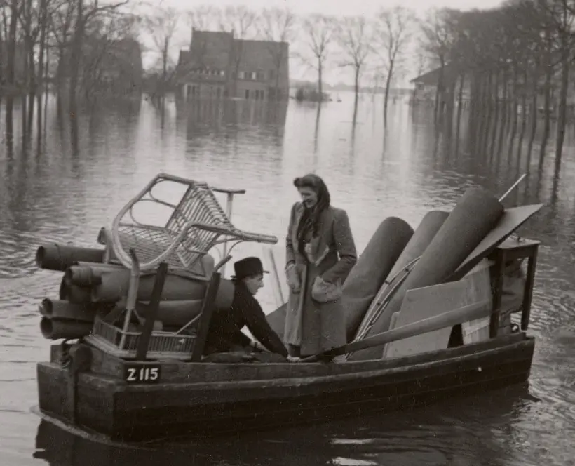Black and white photograph of two people in a boat with stacked furniture, floating in flood waters