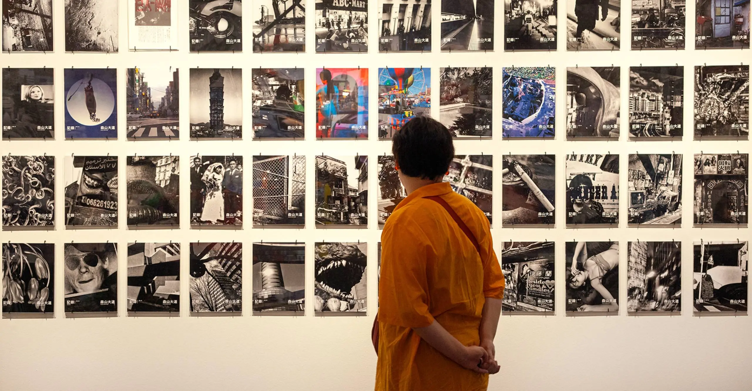 Colour photograph of a person stood infront of a wall of magazine covers by Daido Moriyama